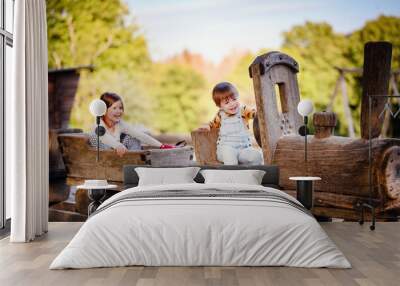 Two children, a boy in overalls and a girl in a sweater, play on a wooden train structure in an outdoor playground, laughing and enjoying the autumn day. Wall mural