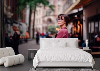 Woman in casual attire and glasses, smiling confidently with her phone and backpack, standing in a bustling city street lined with cafes and people. Wall mural