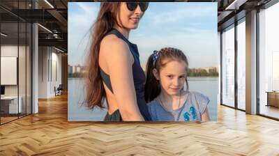 Mother and daughter stand close to each other at the lake shore. The mother smiles, and the girl looks happy. Both are beautiful, of European appearance. High quality photo Wall mural