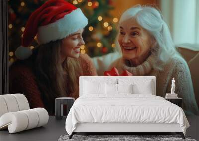 Young girl in Santa hat giving beautifully wrapped gift with red ribbon to elderly woman sitting on sofa with Christmas tree in background, warm relationship between two generations Wall mural