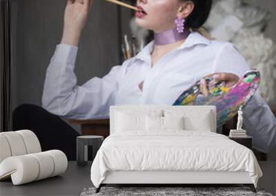 Beautiful young brunette girl sitting on a chair in a white shirt with a palette for drawing on a gray background Wall mural