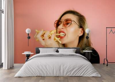 the girl eats French fries on a pink background, the girl originally blows out the French fries like a match, a beautiful young girl eats fast food Wall mural