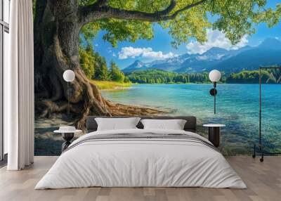 Serene lakeside with a large tree and mountains under a bright blue sky on a sunny day Wall mural