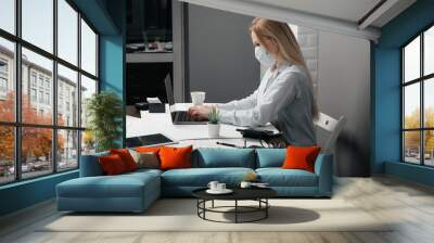 Business girl wearing a mask takes some notes in her laptop while sitting in her office workplace Wall mural