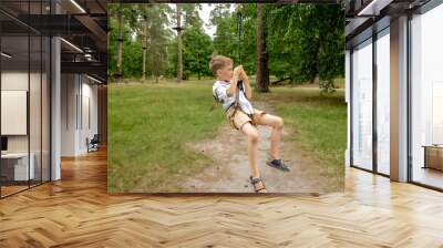 Cheerful laughing boy riding on the zip-line strung between two trees in adventure park. Kids sports, summer holiday, fun outdoors, scouts. Wall mural