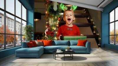 Happy laughing toddler boy holding Christmas gift box from Santa and smiling in camera while sitting on wooden stairs at home. Family celebrations on winter holidays. Wall mural