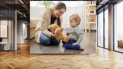 Happy smiling mother playing with her baby son with teddy bear on floor in living room. Wall mural