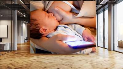 Portrait of cute little baby boy sucking mother's breast. Young woman using smartphone while feeding her newborn child. Concept of healthy and natural baby nutrition. Health of mother and child Wall mural