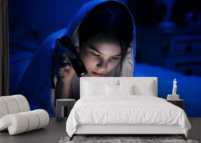 Portrait of teenage girl reading book in bed with flashlight Wall mural