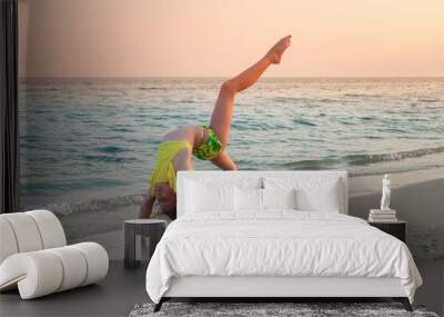 The child does a gymnastic exercise on the seashore. Girl doing back stretching on the sandy beach Wall mural