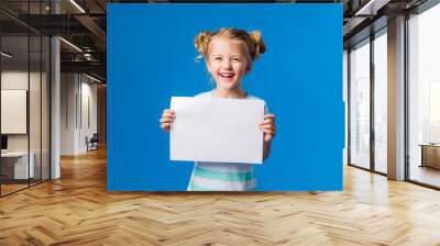 baby girl holding a white sheet.Cute little girl with a white sheet of paper.blue background.space for text.A little girl holds an empty piece of paper Wall mural