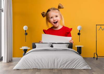 baby girl holding white sheet.Cute little girl with white sheet of paper.yellow background.copy spase.Little girl holding empty sheet of a paper Wall mural