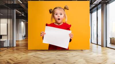 baby girl holding white sheet.Cute little girl with white sheet of paper.yellow background.copy spase.Little girl holding empty sheet of a paper Wall mural