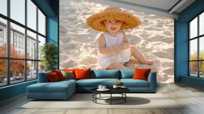 baby girl in white clothes and a straw hat sits on the white sand on the beach in summer Wall mural