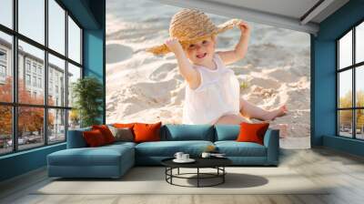 baby girl in white clothes and a straw hat sits on the white sand on the beach in summer Wall mural