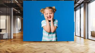 Cute little girl blonde smiling holding hands on sides standing on a blue background, happy child on a blue background, space for text Wall mural