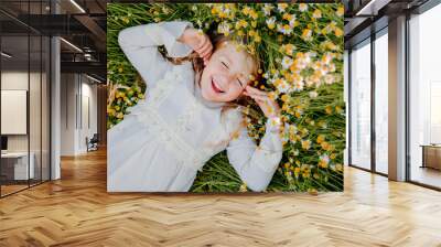 happy little girl in a cotton dress lies in a field of daisies in the summer at sunset. laughs, view from above Wall mural