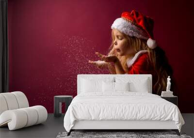 little blonde girl in a Santa suit blows snow off her hands. red background isolate. space for text Wall mural