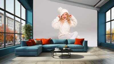 Winter clothes. Portrait of a little curly-haired girl in a knitted white winter hat. little blonde girl in white knitted hat and sweater smiling light background isolate, space for text Wall mural