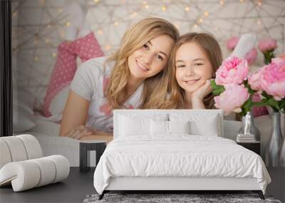 Happy family portrait. Mother and daughter hugging in bed in pajamas and looking at the camera. Loft interior. Background with lights. Pastel colors Wall mural