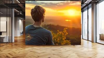 A man sits on a mountain top, looking out at the horizon. The sky is a beautiful mix of orange and pink hues, creating a serene and peaceful atmosphere. The man is lost in thought Wall mural