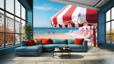 Close up image of a red and white vertical striped beach umbrella on a background of a beach with sun loungers and blue sea Wall mural