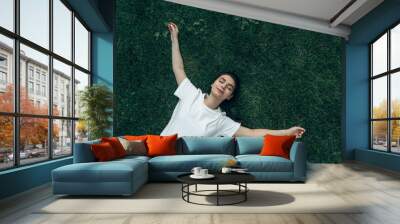 Caucasian girl 20-25 years old lies on the green grass on a summer day smiling, arms outstretched, hugging everyone. View from above. View from the corner. The concept of a happy life, vacation Wall mural