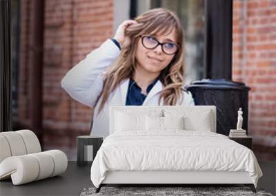 A cute girl in the white jacket with glasses  is adjusting her hair and looking sideways with shy smile while standing outdoors on the background of a brick wall. Dressed in business style Wall mural