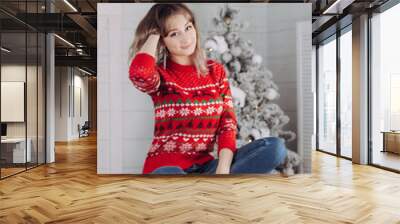 A young caucasian charming smiling woman in a red Christmas sweater with deer and snowflakes pattern siitting on the background of a white fir tree. New Year's interior Wall mural