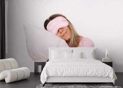 A young caucasian sleepy smiling blonde woman in a pink pajamas and sleep mask on her eyes hugging a pillow isolated on a white background. Insomnia, sweet dreams, cuddling with a pillow Wall mural