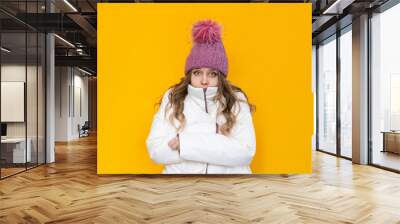 A young frozen caucasian pretty blonde woman in a white warm jacket and pink knitted hat shivers hugging herself isolated on a bright color yellow background. Stylish winter clothes Wall mural