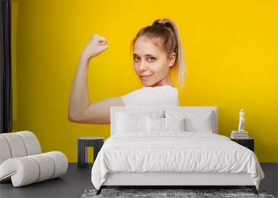 Strong powerful confident caucasian young blonde woman raises arm and shows bicep isolated on a bright colorful yellow background. Feminism, girl power, equal women's rights and independence concept Wall mural