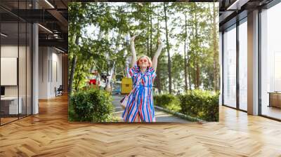 Bright summer image. The girl is blonde in a bright striped shirt dress walking in the amusement Park. Portrait of a woman on vacation. She's fooling around and having fun with the carousel. Wall mural