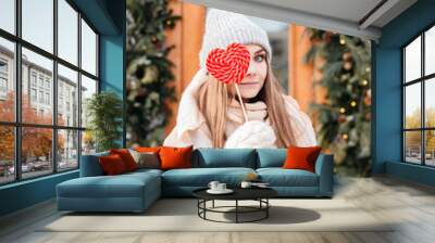 lifestyle portrait of pretty blonde girl walking on the holiday winter city. Woman holding a large Lollipop in the shape of a red heart. Wearing stylish pink coat and wide scarf. Christmas mood Wall mural