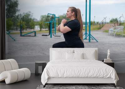 A beautiful fat girl in a black tracksuit is engaged in fitness on the sports ground. Young woman lunges outdoors on a warm summer day. Healthy lifestyle and weight loss. Wall mural