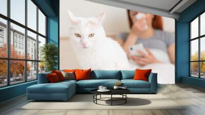 A woman suffering from an attack of an allergy blows her nose in her nose raft. White cat in the foreground. Girl with a cold in bed at home. Wall mural