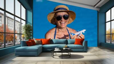 An elderly woman in a hat and sunglasses holds a model airplane while sitting by the pool. Retirement vacation concept.  Wall mural