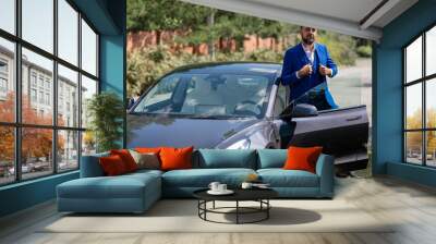 Caucasian bearded man in a blue suit gets out of a black electro car in the countryside in summer. Wall mural