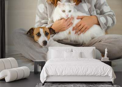 Caucasian woman holding a white fluffy cat and Jack Russell Terrier dog while sitting on the bed. The red-haired girl hugs with pets. Wall mural