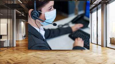 Close-up of a female receptionist wearing a facial mask talking on a headset while sitting at a work desk in the office. Portrait of a manager working during a coronavirus epidemic. Wall mural