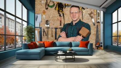 Portrait of a carpenter in goggles and overalls holding a wooden hammer in the workshop against the background of a wall with tools. Wall mural