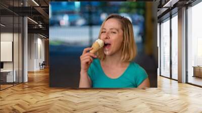 Portrait of a cute girl in a green dress is walking outside and eating dessert. Beautiful blonde enjoying a cone with ice cream on a beautiful summer day. Life is a pleasure Wall mural