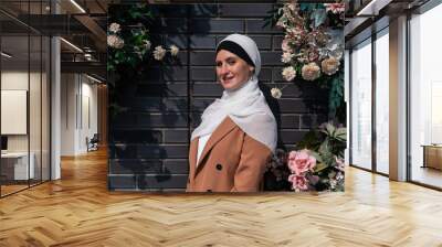 Portrait of a young caucasian woman dressed in a hijab near a wall with flowers. Wall mural