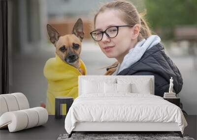 Portrait of a young Caucasian woman hugging a small dog in a jacket.  Wall mural