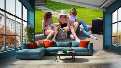 Three Caucasian women and a dog go on a trip. The girls are sitting on the curb with suitcases and waiting for a taxi. Summer vacation concept together with girlfriends Wall mural