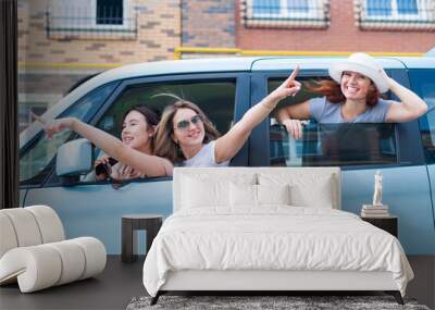 Three joyful Caucasian girlfriends look out of the car window. Excited women go on a road trip. The brunette blonde and the redhead are going on vacation. Wall mural