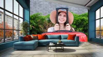 Young beautiful caucasian woman in a hat looks out of a red suitcase with a handle. Happy girl goes on a trip with luggage. Summer vacation concept. Wall mural