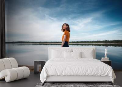 Red-haired girl in an orange T-shirt and orange glasses on the shore of a lake  Wall mural