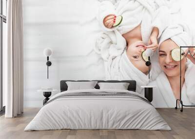 Positive mother and daughter doing face mask applying pieces of cucumber to their eyes, wrapped in a white bath towel, Facial skin care, cosmetology and spa, Long banner format. space for text Wall mural