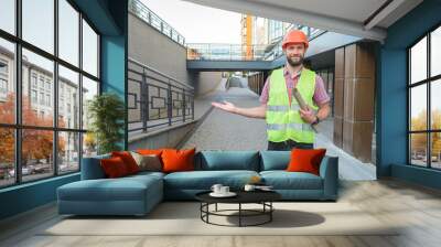 Male architect in orange helmet holding blueprint and presenting empty hand
 Wall mural